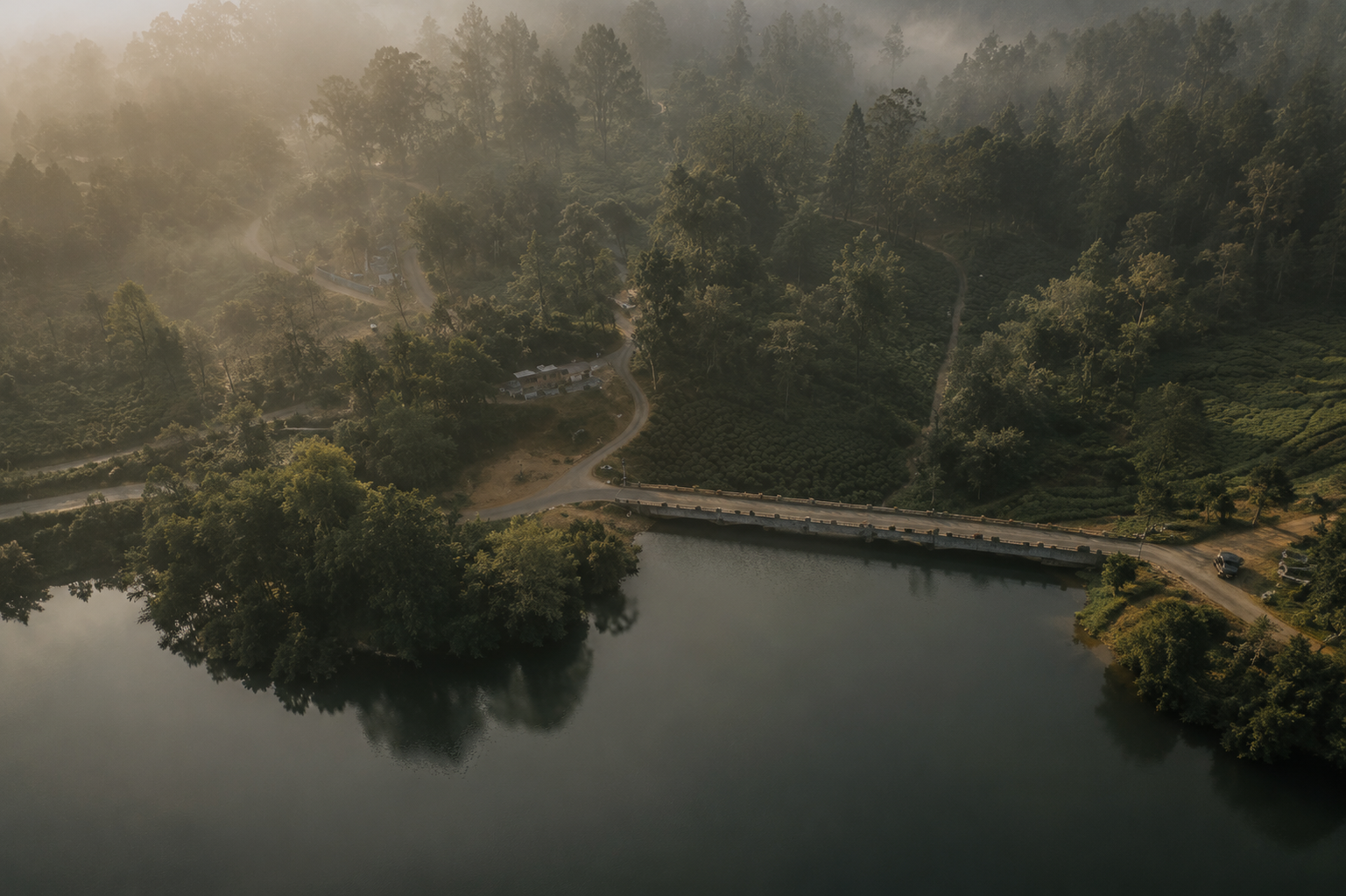 Aerial view of a lake and small dam bridge surrounded by misty forest and tea plantations